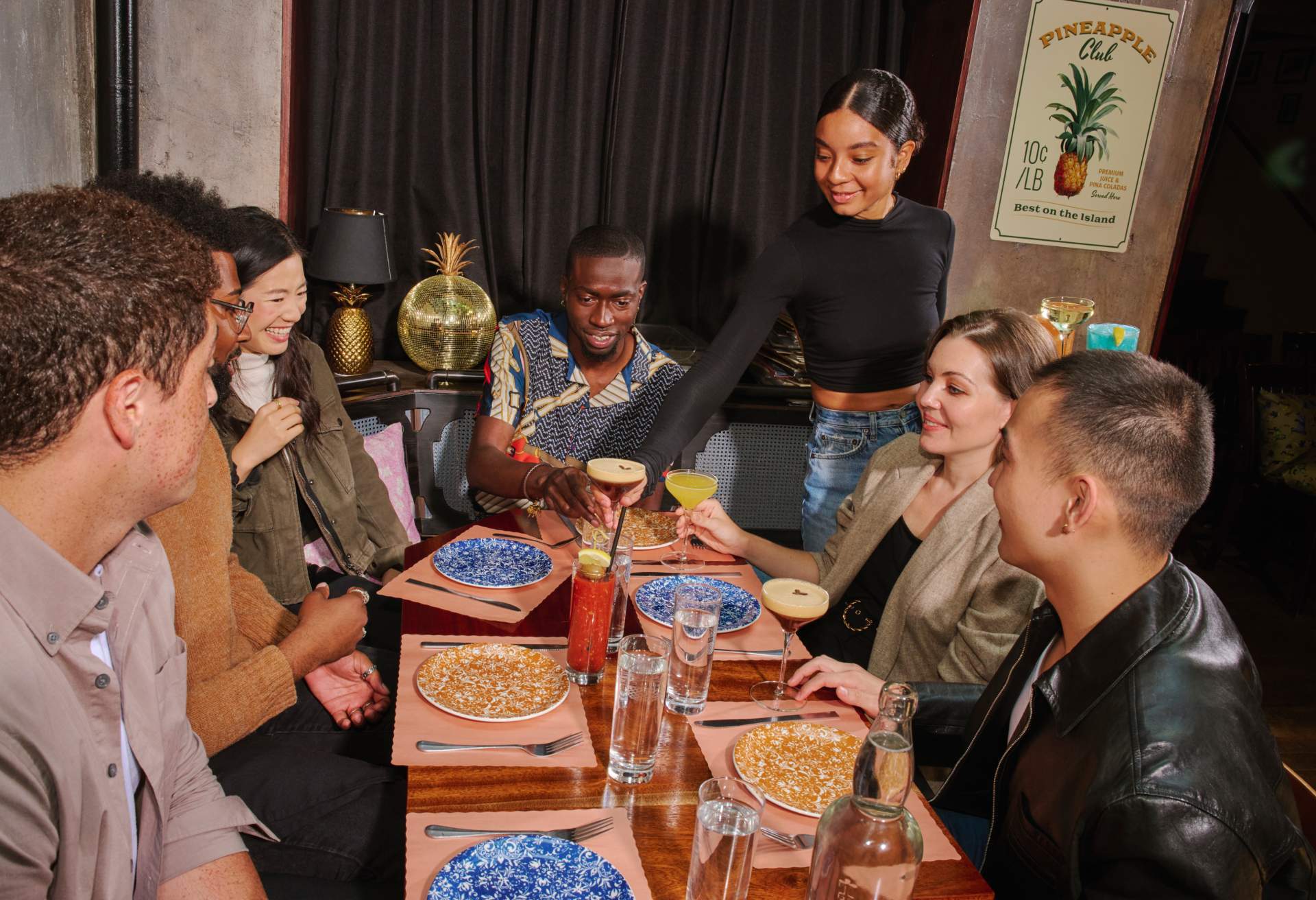 Das Bild zeigt eine große Gruppe von Gästen, die an einem Restauranttisch sitzen, während eine Kellnerin ihnen Getränke serviert