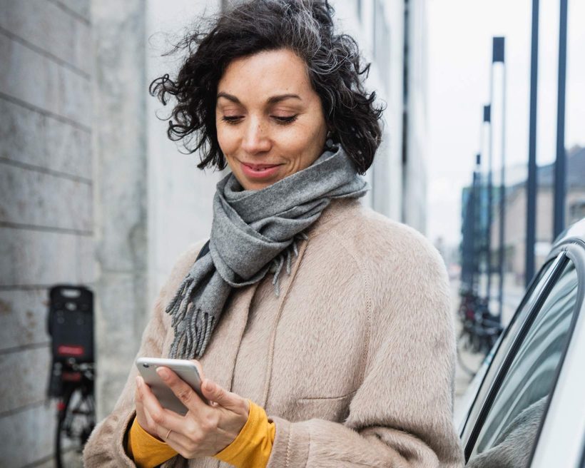 Die Abbildung zeigt eine Person, die draußen ein Smartphone hält. Die Person trägt einen Mantel und einen Schal. Sie steht neben einem Auto. Die Person schaut auf das Gerät, das sie in den Händen hält.
