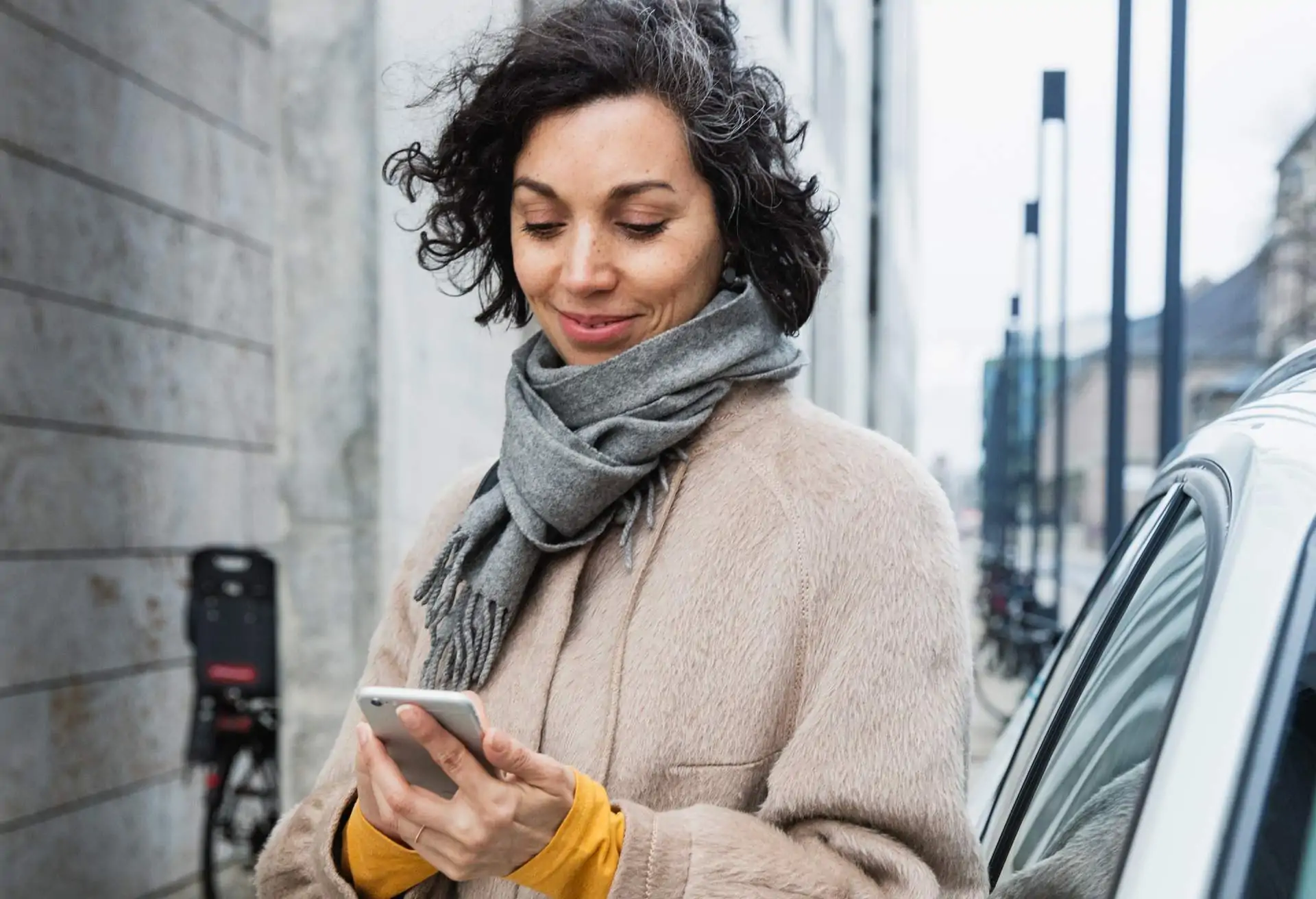 Die Abbildung zeigt eine Person, die draußen ein Smartphone hält. Die Person trägt einen Mantel und einen Schal. Sie steht neben einem Auto. Die Person schaut auf das Gerät, das sie in den Händen hält.