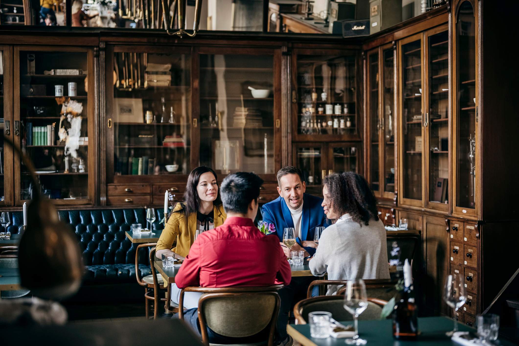 Die Abbildung zeigt vier Gäste, die gemeinsam an einem Tisch sitzen. Das Restaurant ist im Stil eines altmodischen Büros eingerichtet. Mit Leder überzogene Sitzbänke und Bücherregale sind in die Wände eingebaut. Die Gäste tragen Kleidung in unterschiedlichen Farben. Sie unterhalten sich miteinander.