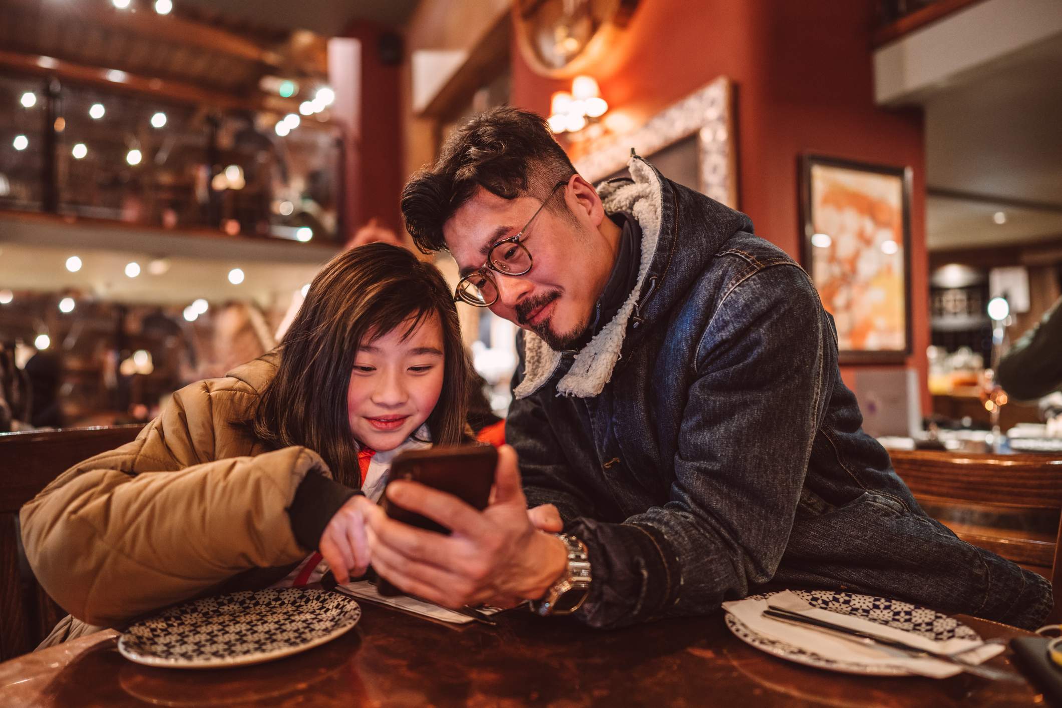 Die Abbildung zeigt ein Kind und seine Eltern, die gemeinsam ein Smartphone benutzen, während sie an einem Tisch in einem Restaurant sitzen. 