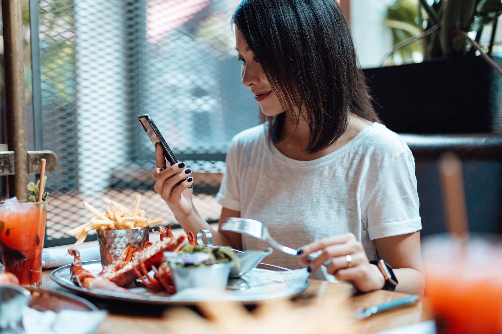 Die Abbildung zeigt eine Frau in einem Restaurant, die lächelt, während sie auf ihr Smartphone sieht. Vor ihr befindet sich ein farbenreiches Getränk und ein Teller mit Meeresfrüchten und Pommes. 