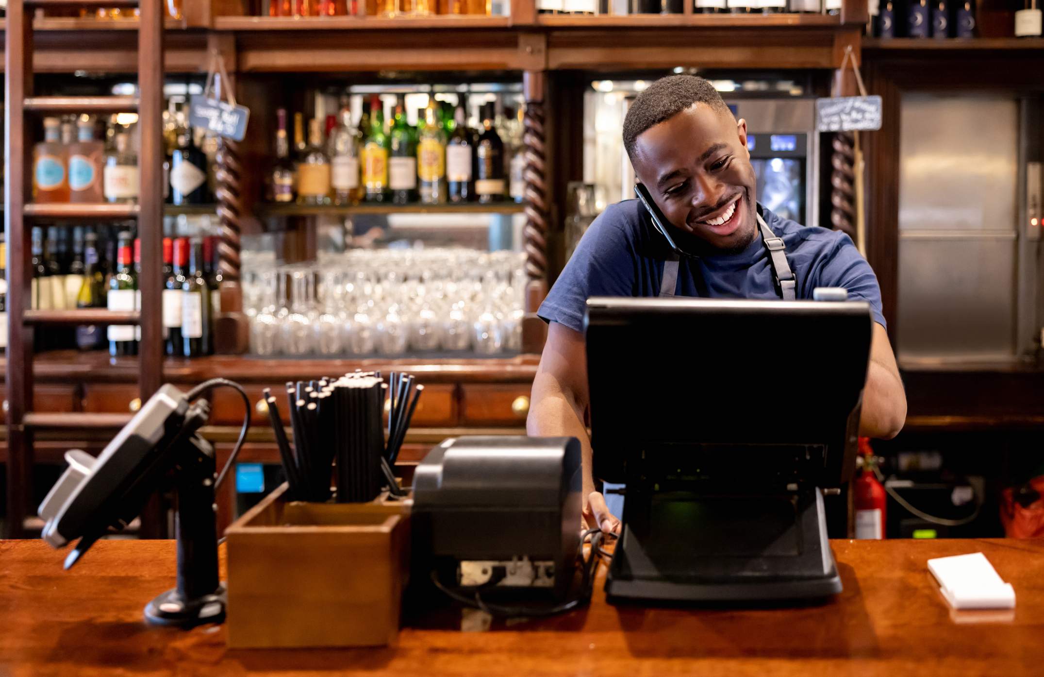 Bild eines Kellners mit blauem T-Shirt und schwarzer Schürze, der telefoniert und gleichzeitig an einem vor ihm stehenden Computer arbeitet. Hinter ihm befindet sich eine Bar. 