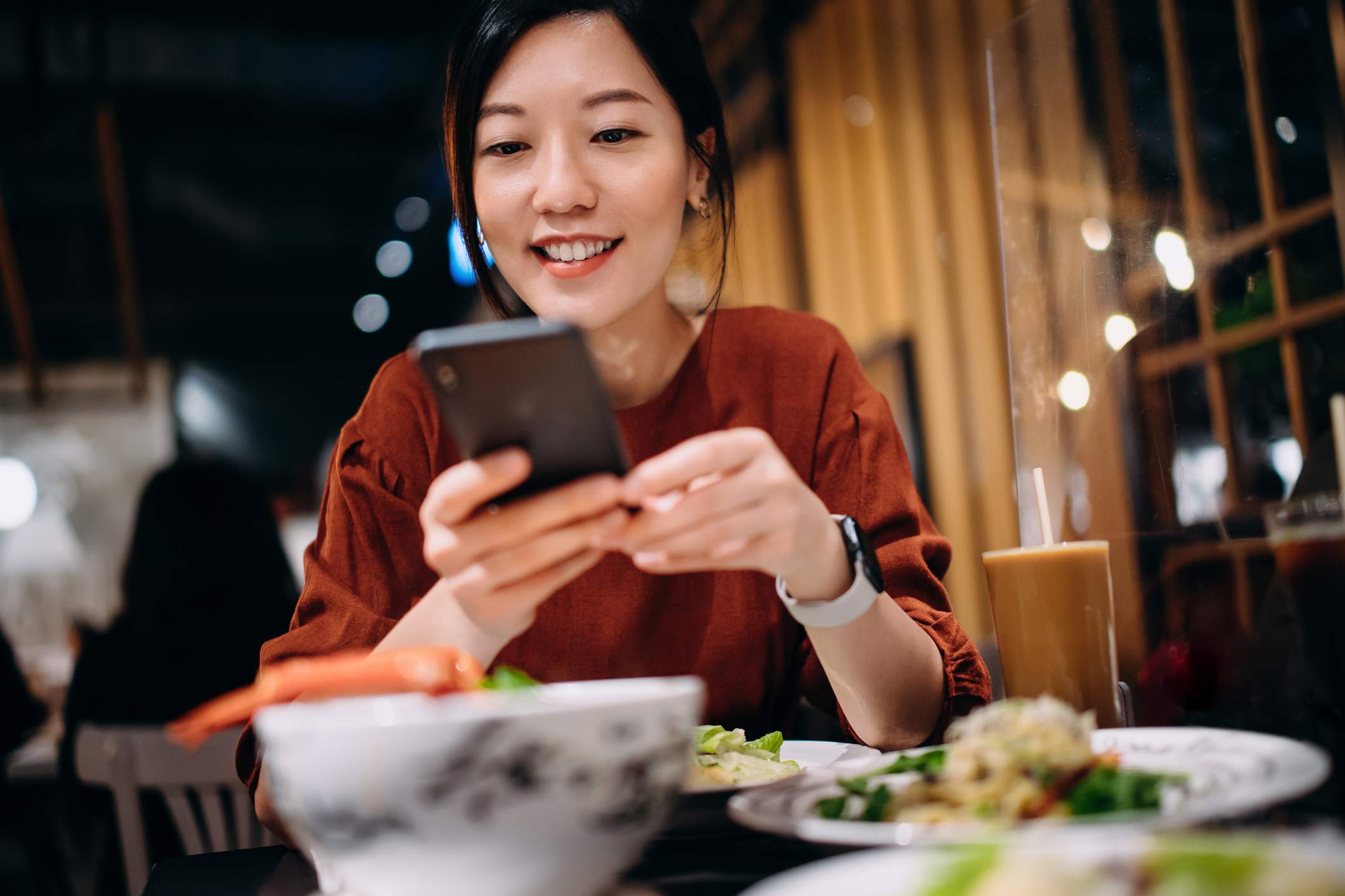 Das Bild zeigt eine Kundin in einem Restaurant, die ihr Telefon benutzt. Sie trägt ein rotes Hemd und eine Smartwatch und lächelt, während sie ihr Telefon hält.
