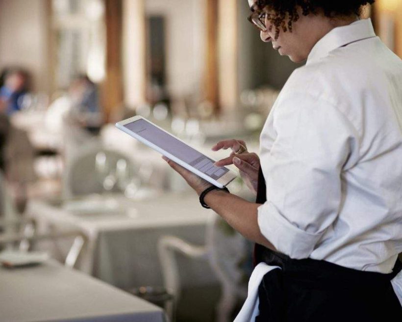 Das Bild zeigt eine Kellnerin, die ein weißes Hemd und eine schwarze Schürze trägt. Sie tippt etwas auf der Tastatur ihres Tablets.