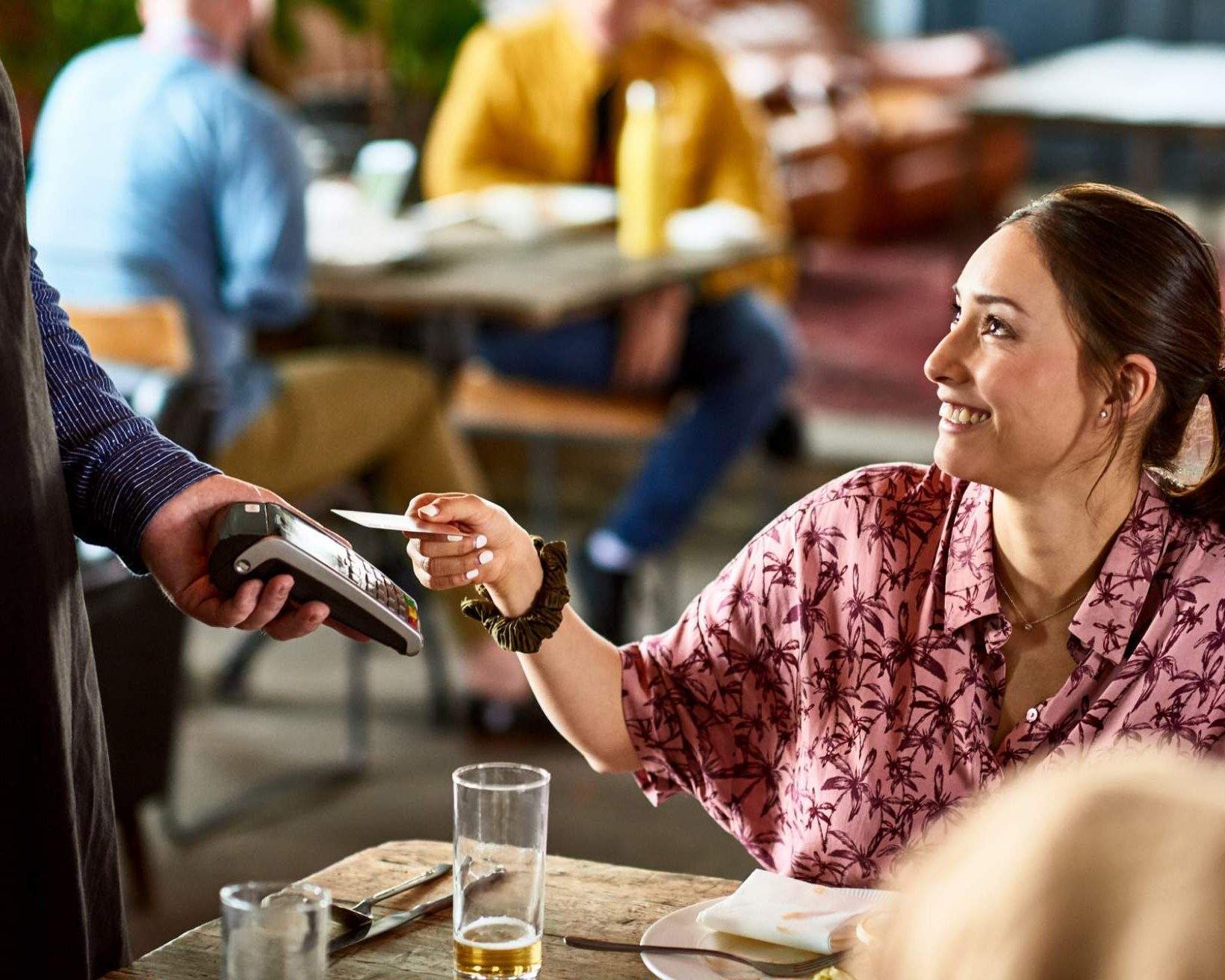 In dieser Abbildung sind ein Gast und ein Kellner in einem Restaurant zu sehen. Der Kellner hält ein Gerät mit einem Zahlungssystem und der Gast legt eine Karte auf das Gerät.