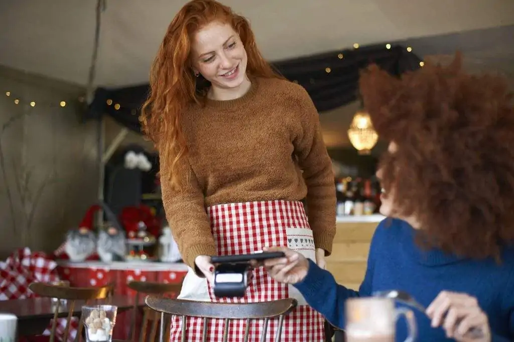 Das Bild zeigt zwei Personen in einem Café. Eine Person ist ein Gast, die andere Person ist eine Mitarbeiterin. Der Gast verwendet das digitale Wallt seines Smartphones zum Bezahlen und tippt legt dieses zur kontaktlosen Zahlung an das Gerät.